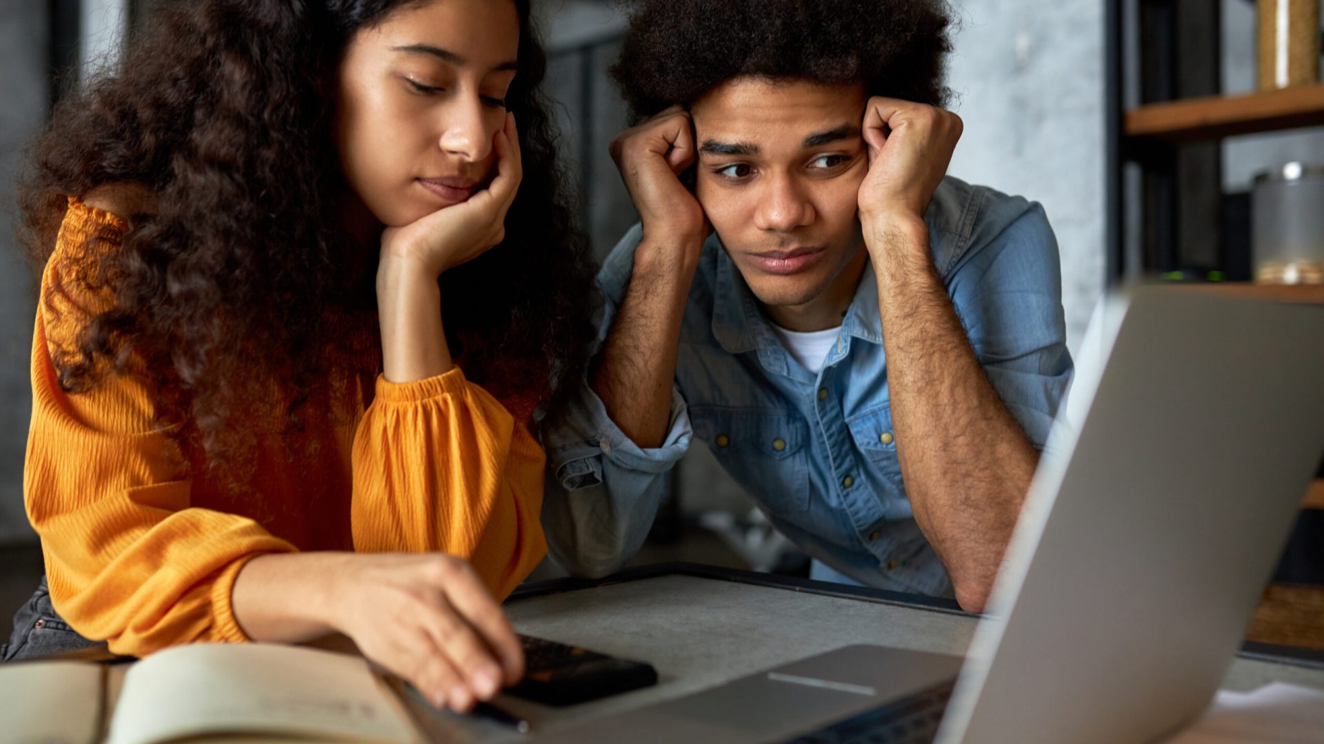 Young married couple of african ethnicity sitting in front of laptop with puzzled facial expressions, man looking at screen holding head in hands, female using calculator, counting expenses