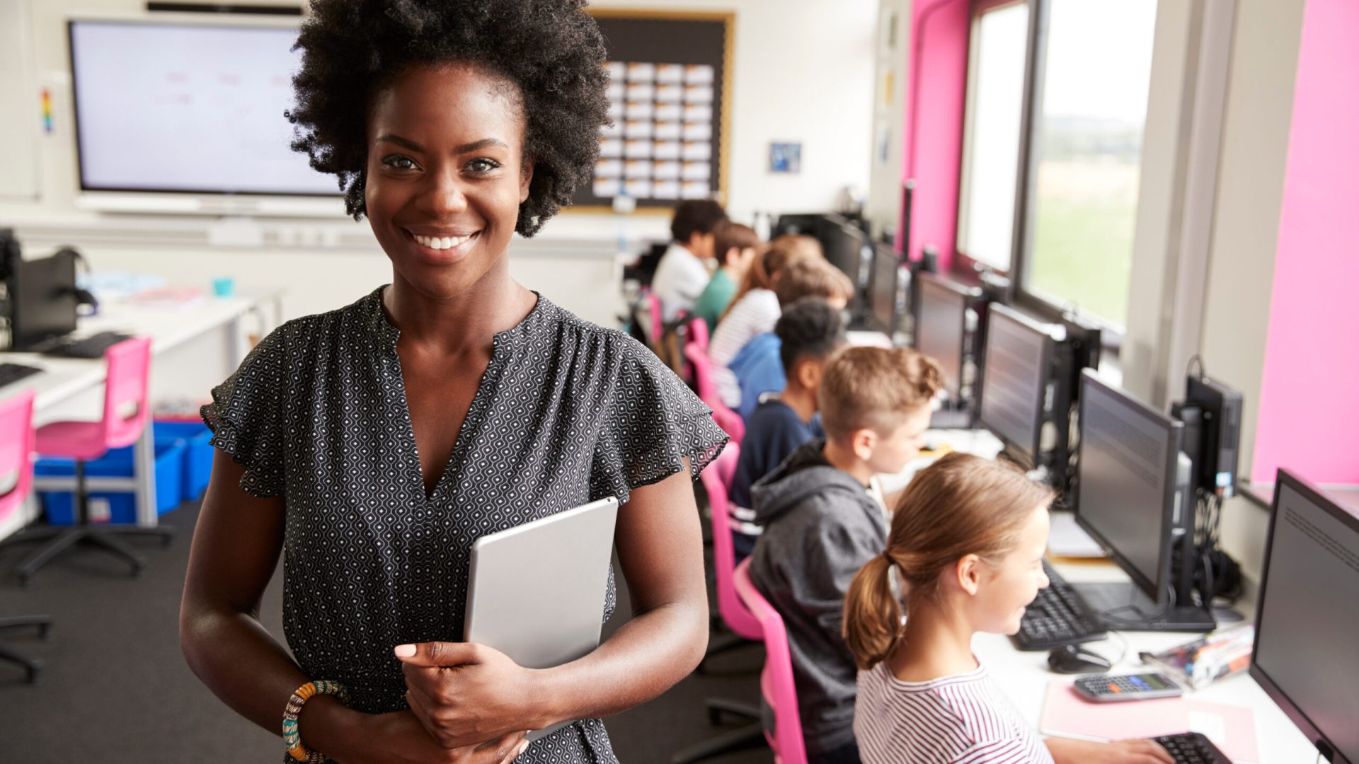 Portrait Of Female Teacher Holding Digital Tablet Teaching Line Of High School Students Sitting By Screens In Computer Class