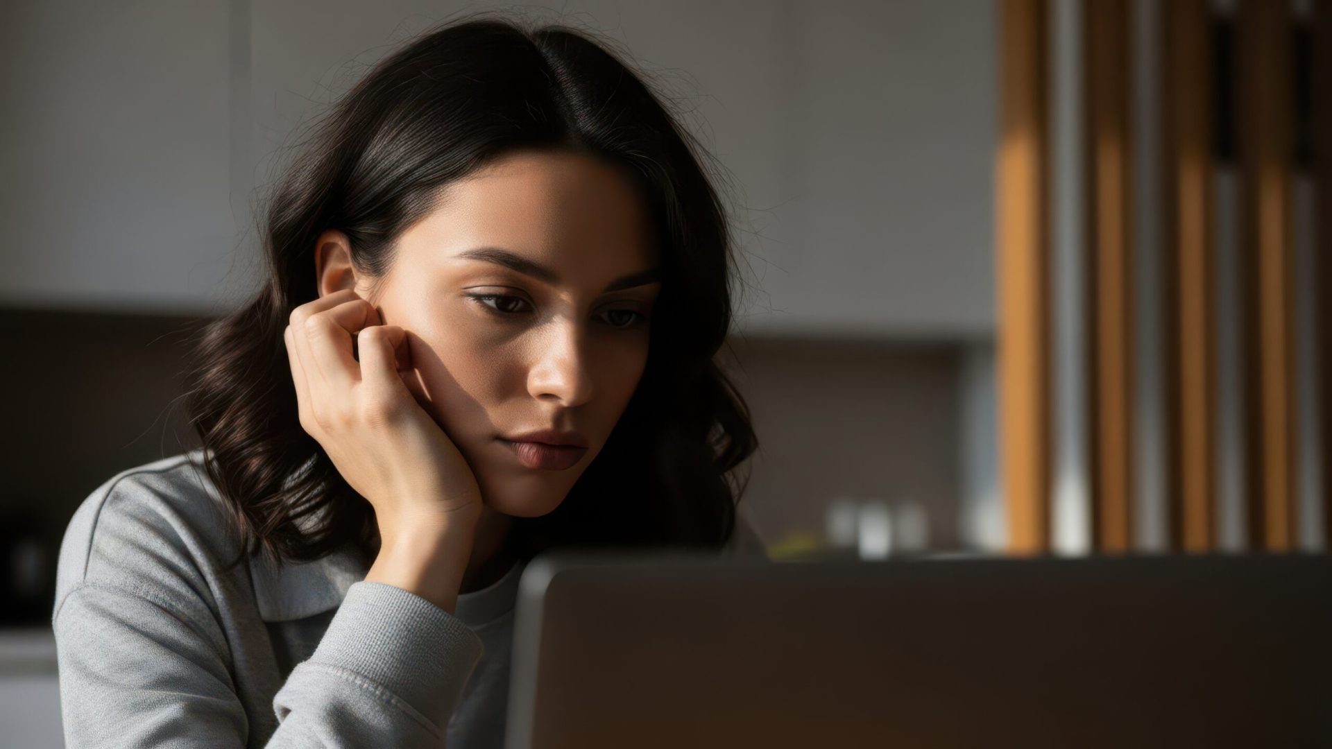 A young woman with dark hair is looking intently at a laptop, her hand resting thoughtfully on her cheek, illuminated by natural light.