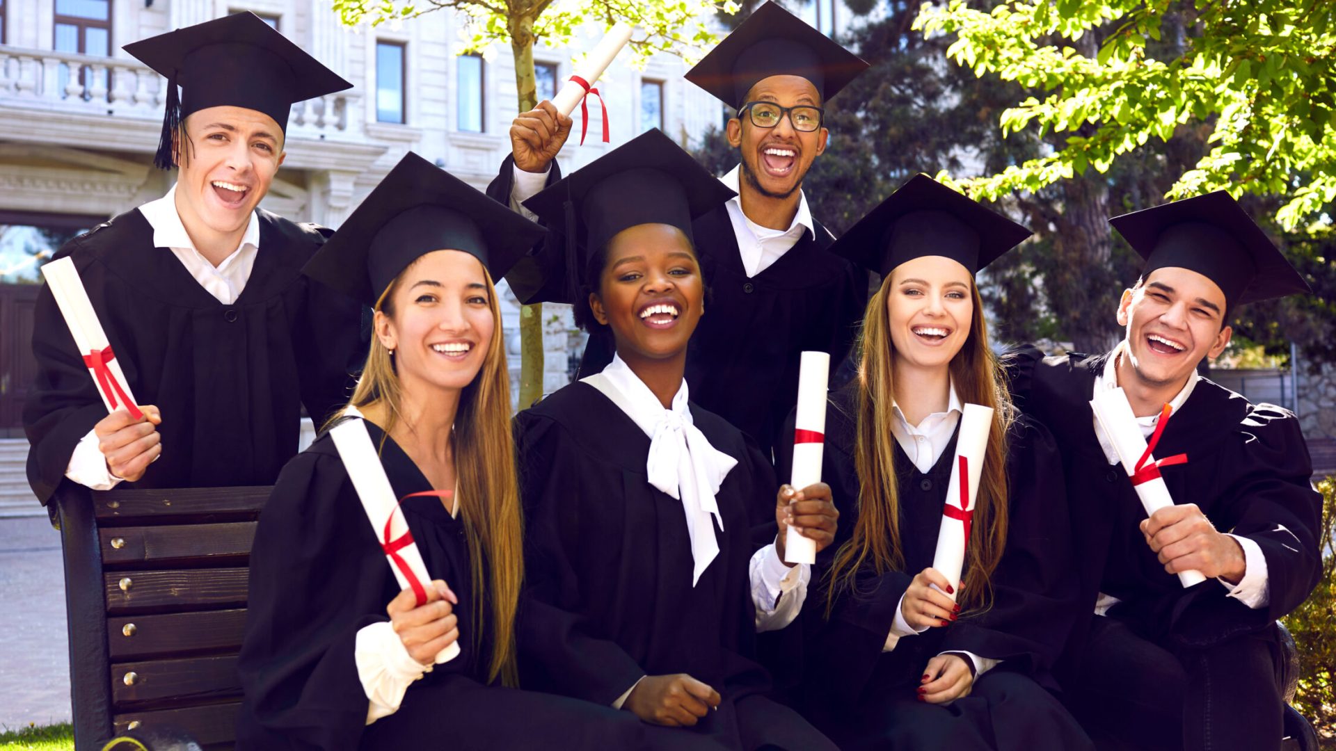 Cheerful diverse university students celebrating graduation outside. Happy graduates in caps and gowns sitting on bench in green campus yard, looking at camera and smiling. Group portrait. Banner