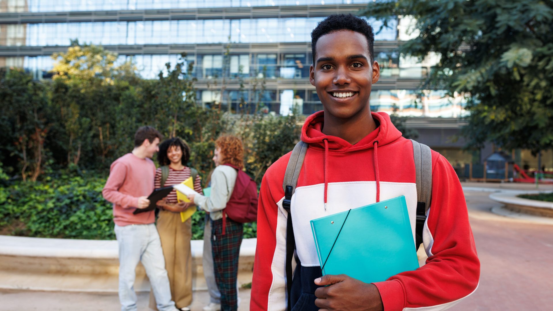 Portrait of a happy african american university student holding a binder on campus with friends talking in background