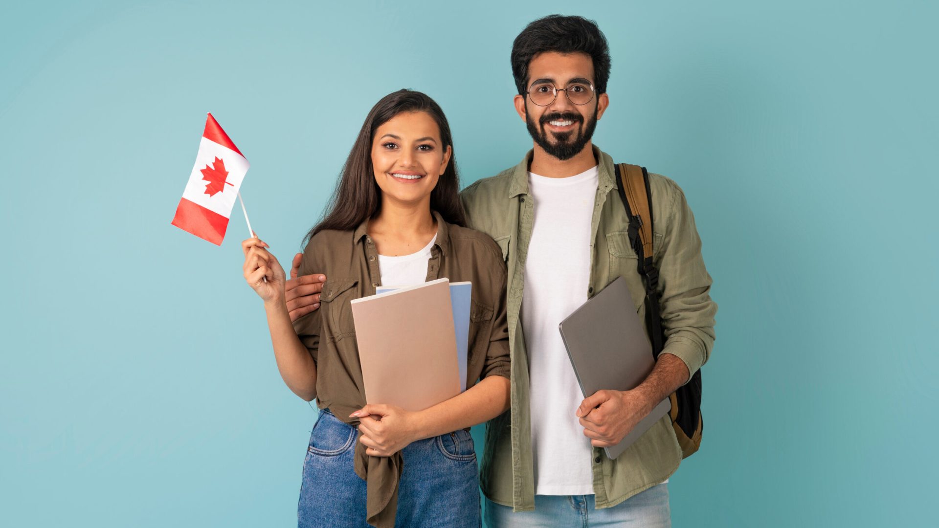 Education abroad, immigration to EU, language school. Cheerful multicultural young arab man and indian woman students with Canadian flag, books and laptop on blue background