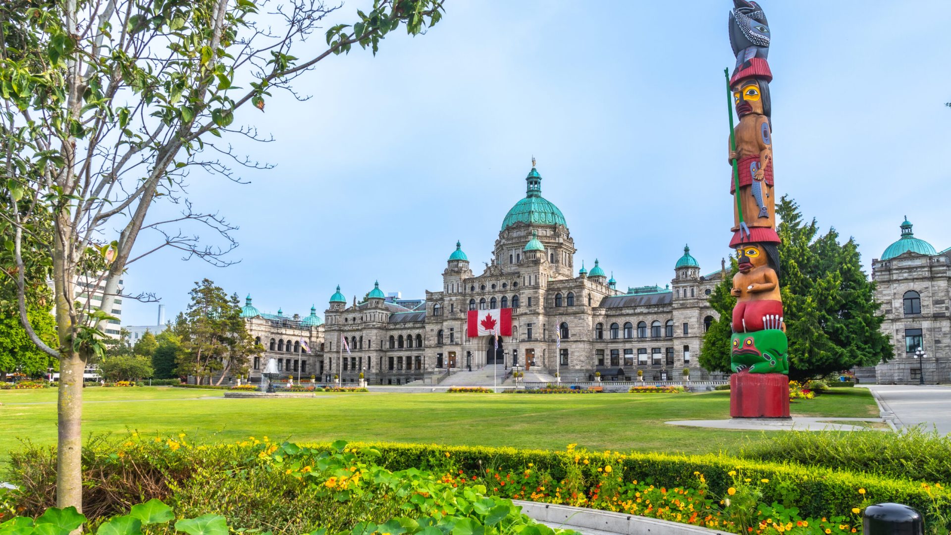 Canadian flag waving in the wind in front of the british columbia parliament buildings with a colorful totem pole in the foreground, located in victoria on vancouver island