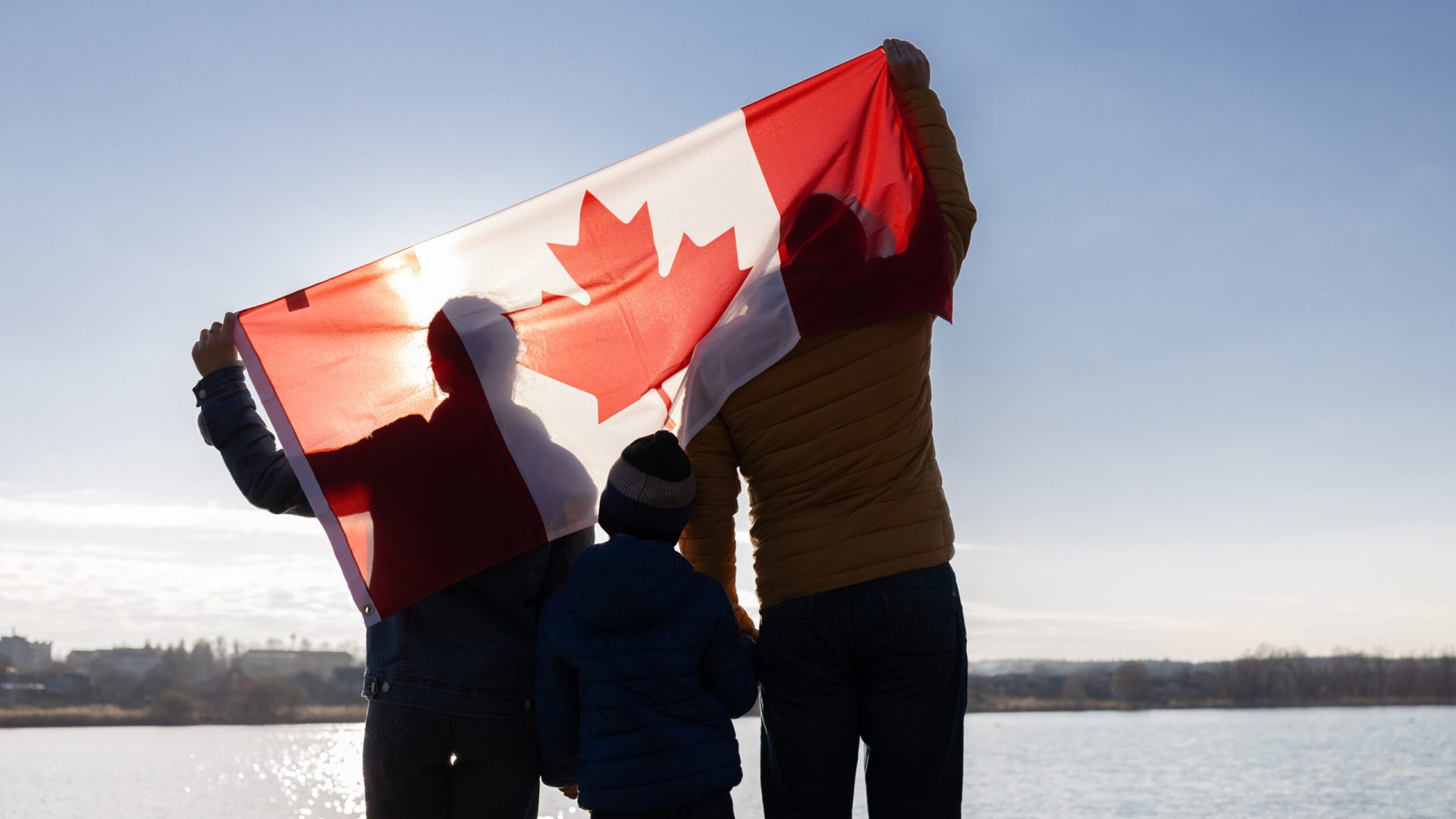 the family holds the Canadian flag behind their backs against the background of the sky and river. Travel, immigration. Happy Canada Day. Pride, freedom, national symbol.