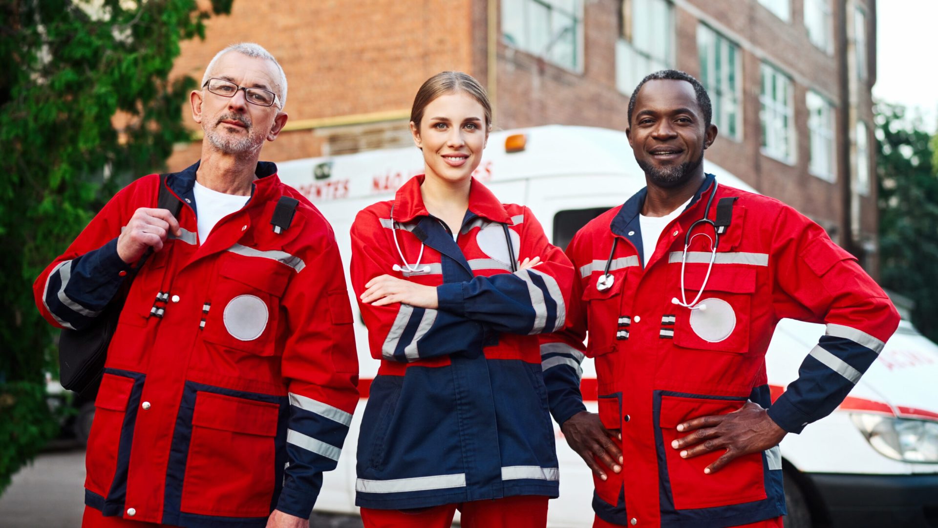 Portrait of team of paramedics in red medical uniform posing outdoors. Male and female doctors standing in street near ambulance and smiling at camera. Medical workers. Professional nurses, first aid