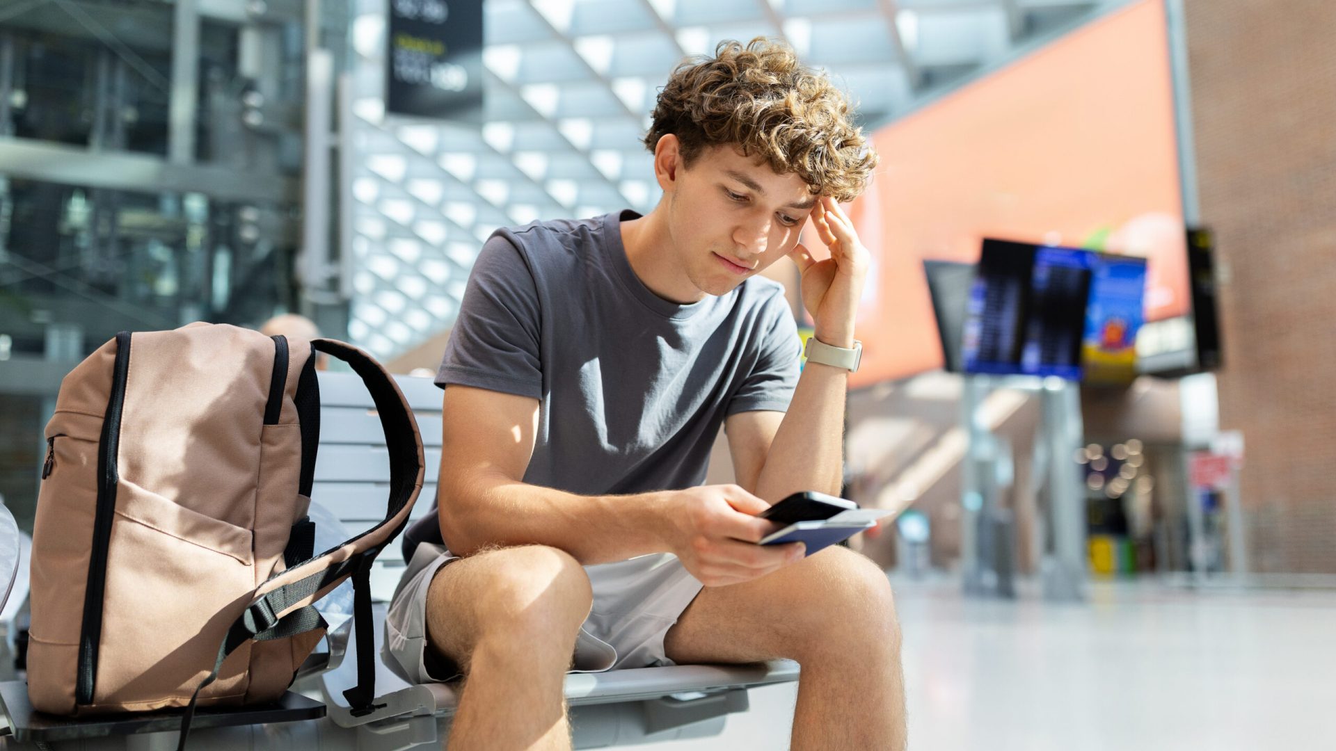 Young man with backpack sitting at airport looking worried at passport and smartphone. Represents stress, flight delay, and challenges of travel lifestyle